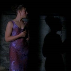Scene in The Sixth Sense. Anna Crowe, played by Olivia Williams, is wearing a purple dress and dolled up with makeup. She's holding a glass of wine in a basement. Her shadown is ominously against the wall.